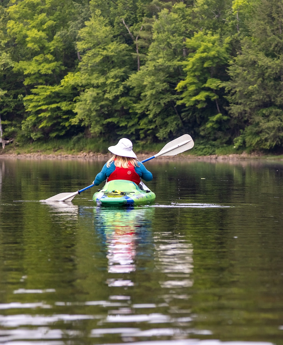 A kayaker wearing a wide-brimmed hat and red life vest paddles across a calm, tree-lined lake, viewed from behind.