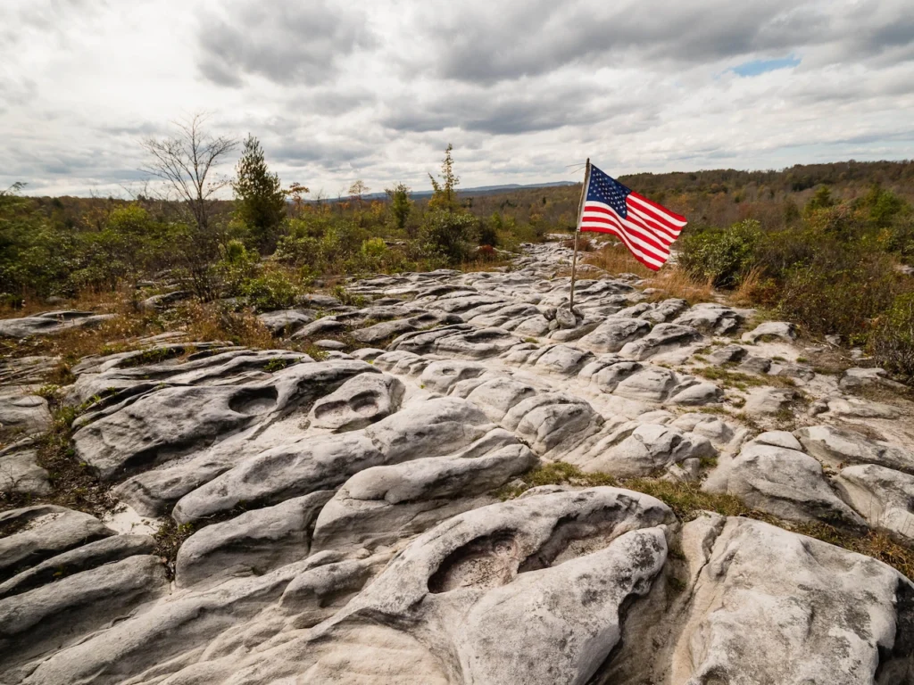 An American flag stands on a pole above a wide stretch of rugged, weathered rock formations under a cloudy sky.