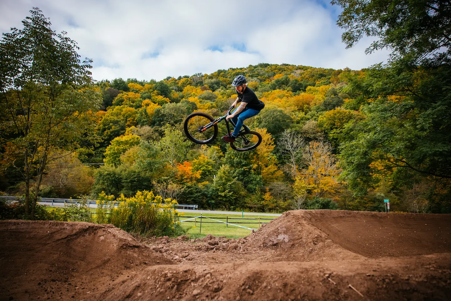 A mountain biker catches air off a dirt jump, soaring above a trail with vibrant autumn foliage covering the hillside behind them.