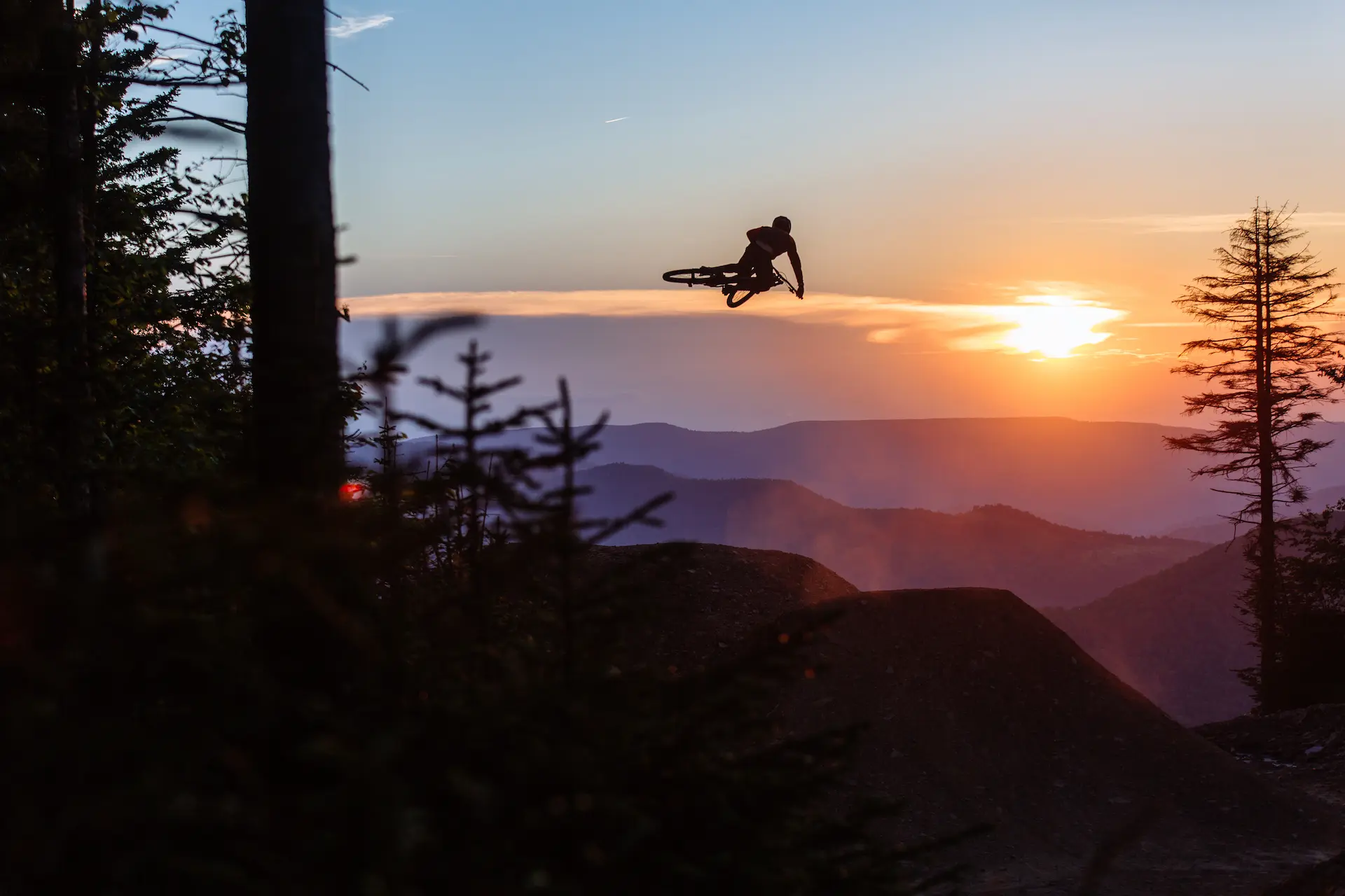 A mountain biker performs a high jump on a dirt trail at sunset, silhouetted against a glowing orange and purple sky over rolling mountain ridges.
