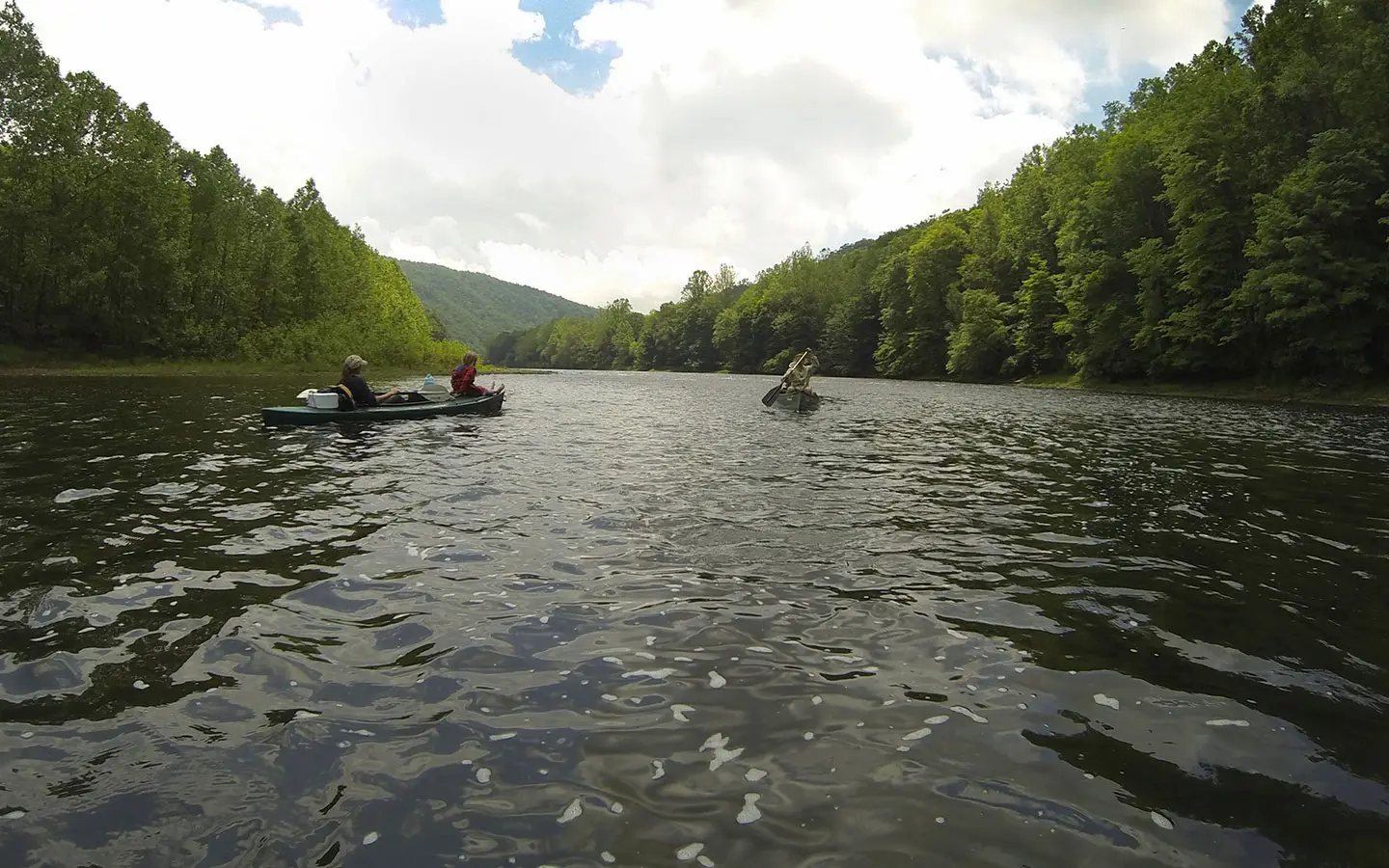 Two people paddle canoes down a wide, calm river surrounded by forested hills under a partly cloudy sky.