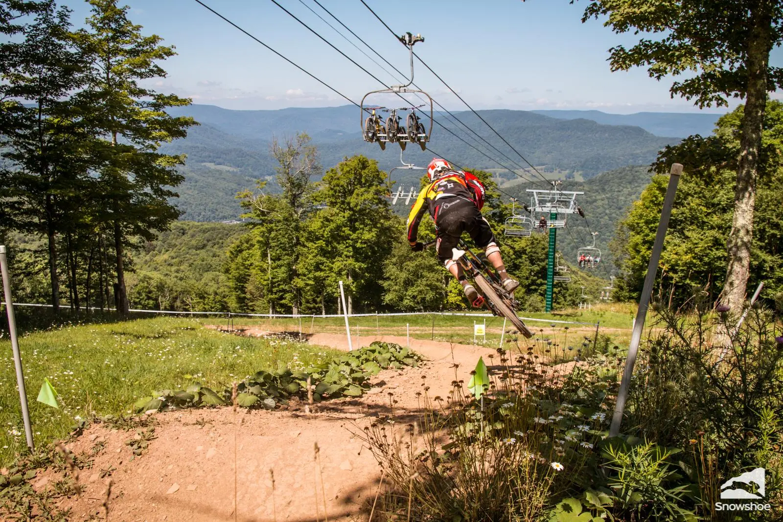 A mountain biker rides downhill over a dirt trail at a bike park, with chairlifts carrying bikes overhead and forested mountains stretching across the background.