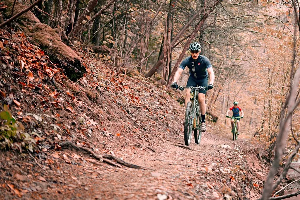 Two mountain bikers ride along a narrow leaf-covered trail through a forest with autumn colors.