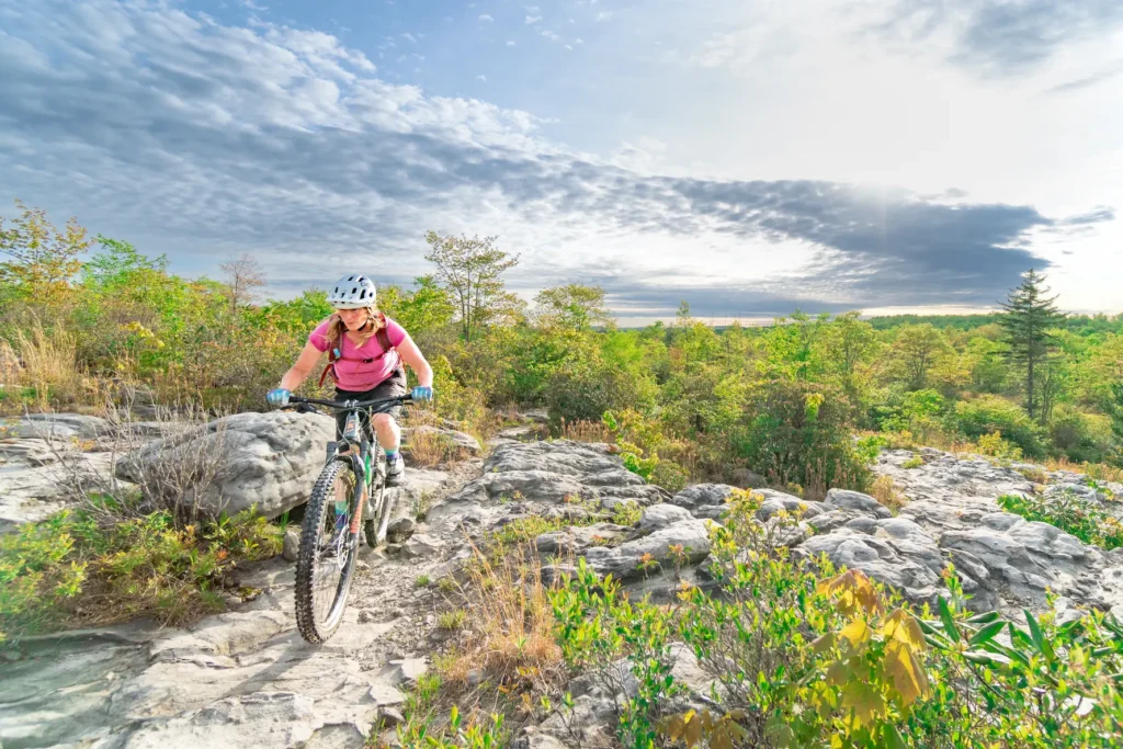 A mountain biker rides over a rocky trail, referred to as the Camp 70 moon rocks, surrounded by bright green foliage, with dramatic clouds and sunlight stretching across the sky in the background.