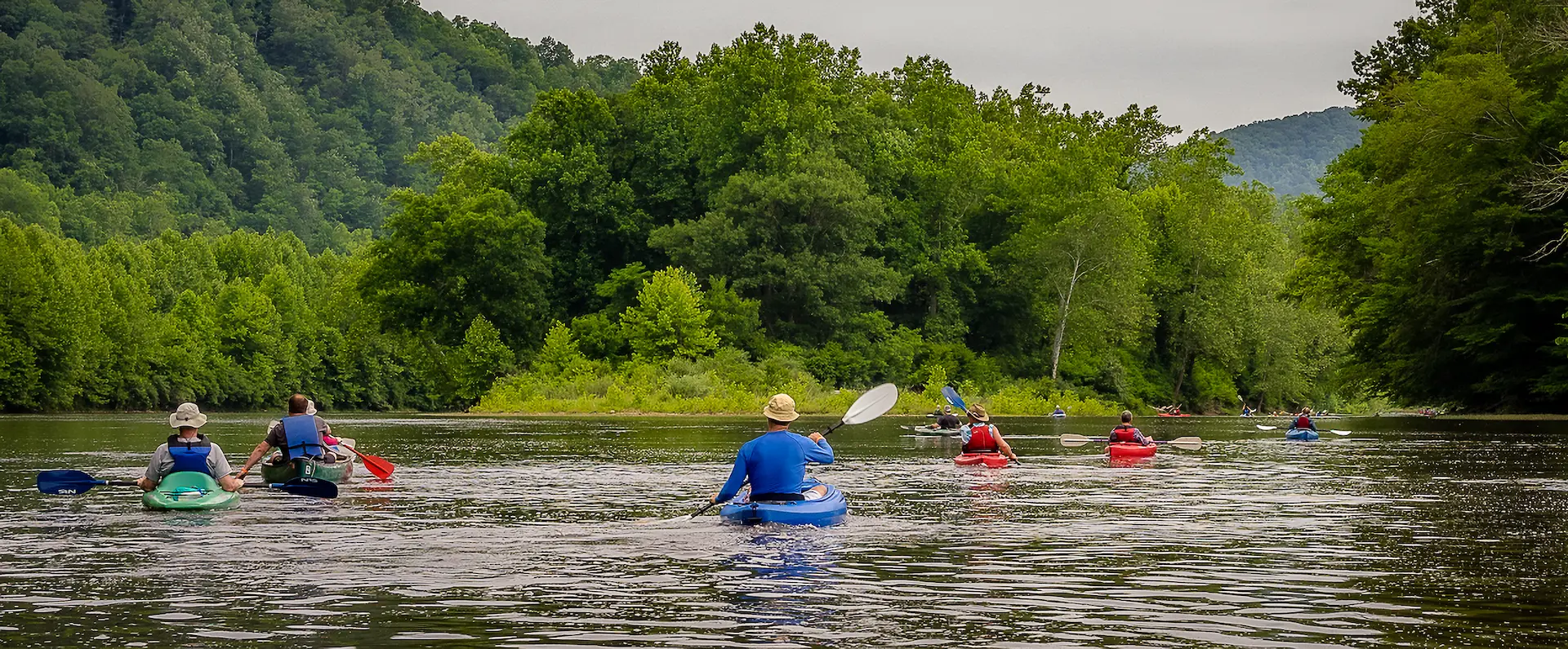 Group of kayakers paddling down wide river surrounded by dense forested hills