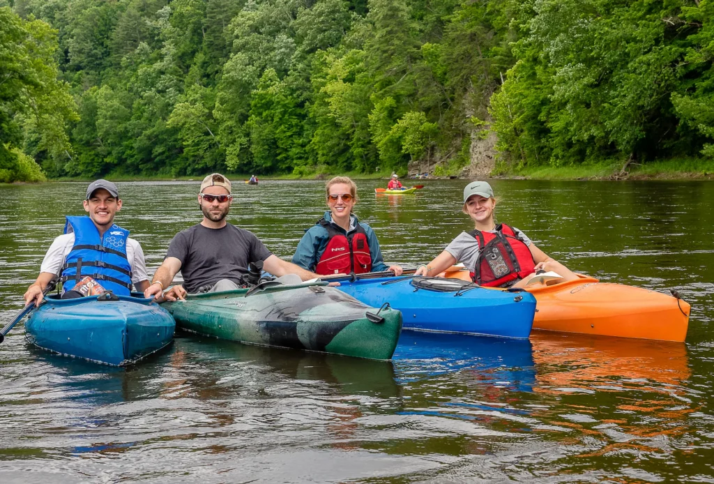 Four adults in kayaks wearing life vests on calm river with wooded shoreline